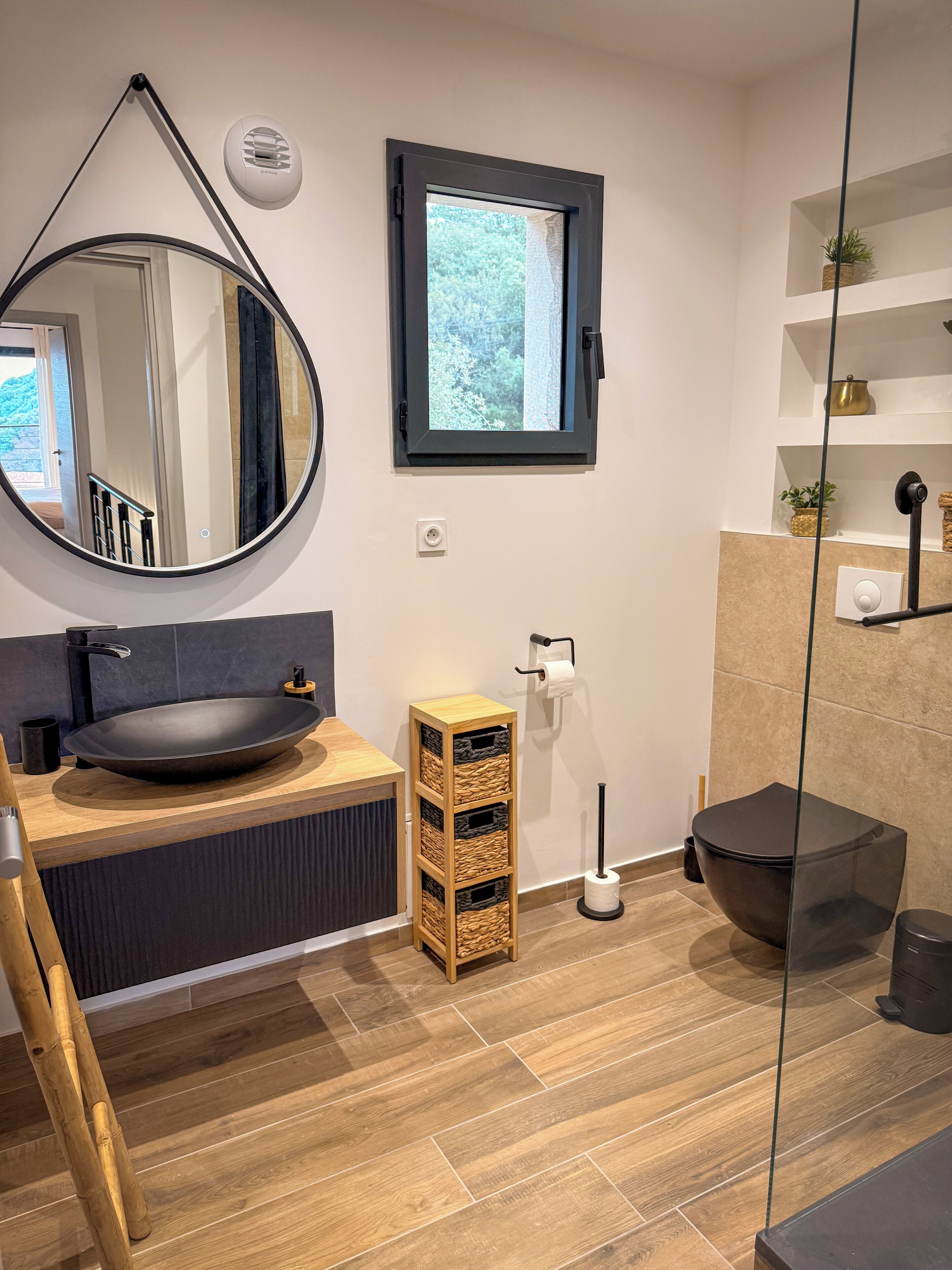 A modern bathroom with a round mirror, a vessel sink, a toilet, and a window with a view of greenery.
