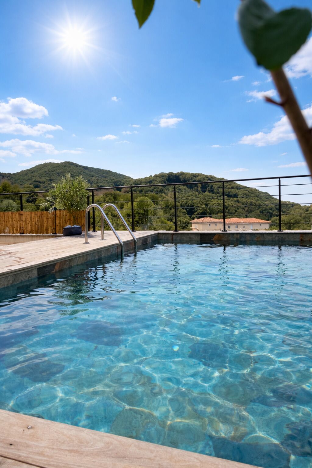 Ein klares Schwimmbecken mit Holzterrasse, Metallleiter und Blick auf einen Berg sowie einen Wald im Hintergrund.