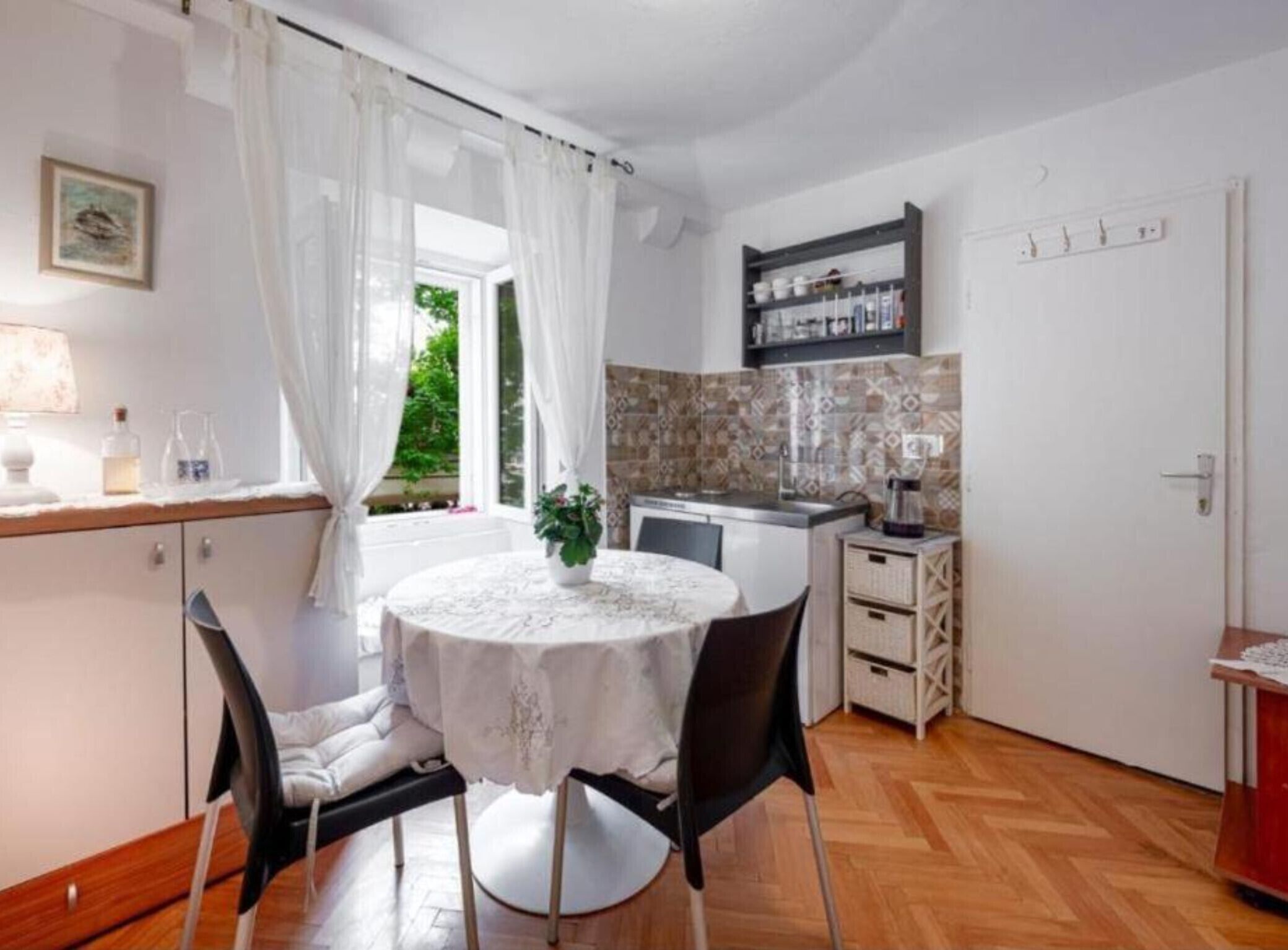 A compact kitchen with a dining area, a window with curtains, and a small plant on the table.