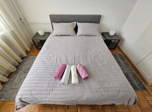 A neatly made bed with a grey bedspread, two pillows, and three rolled towels in pink, white, and grey. - Apartman FITT - Apartman (Zagreb)
