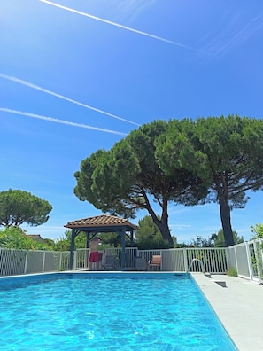 A swimming pool with a pergola and lounge chairs, surrounded by trees and a clear sky.