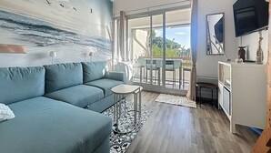 Una sala de estar con un sofá azul, una mesa de centro, un tapete estampado, un cuadro de pájaros y un balcón con vistas.