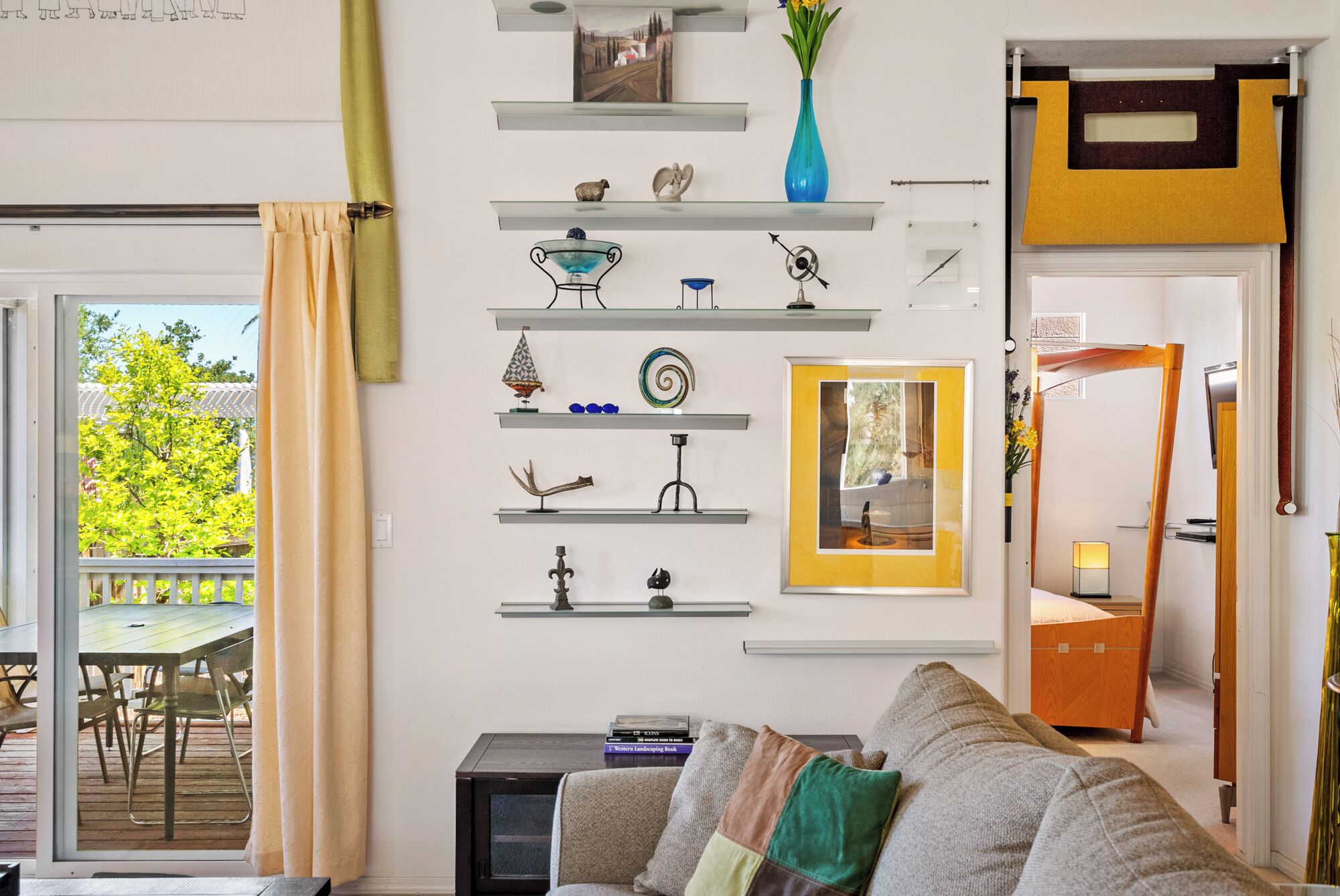 A living room with a sofa, a coffee table, and a TV stand. There are shelves with various decorative items and a window with a view of greenery.