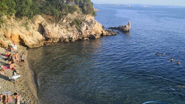 Ein Küstenstrand mit felsigem Gelände, klarem Wasser und ein paar badenden Personen.