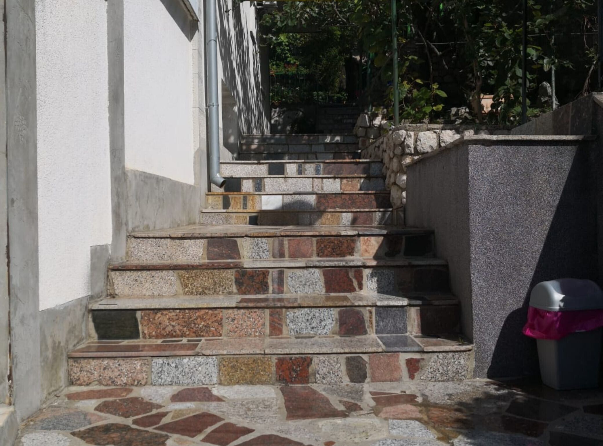 A set of stone steps leading up to a building entrance, with a trash bin and a chair nearby.