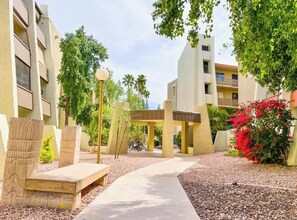 A pathway leading to a covered area with benches, surrounded by buildings and greenery.