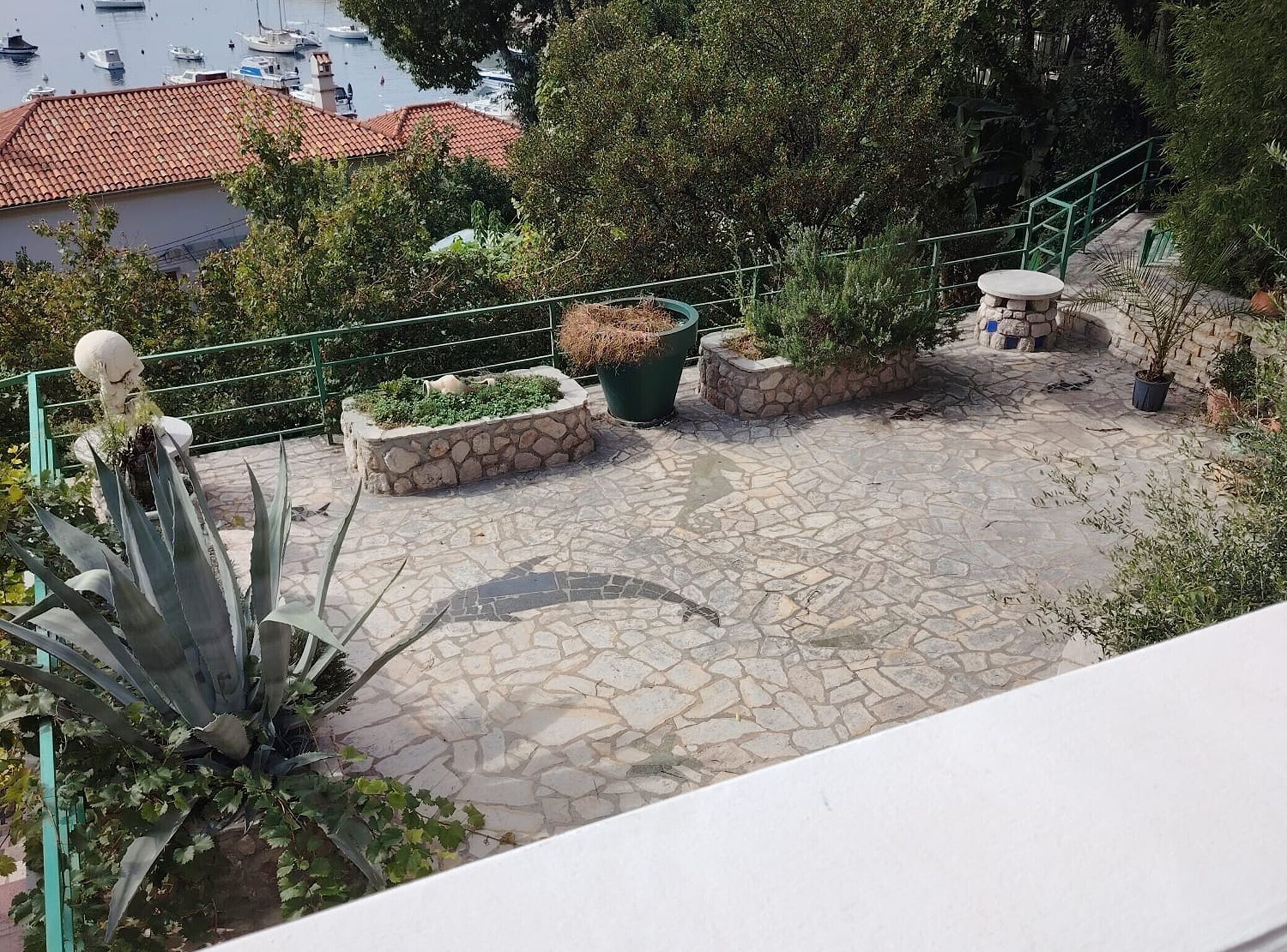 A stone patio with planters, a railing, and a view of boats in the background.