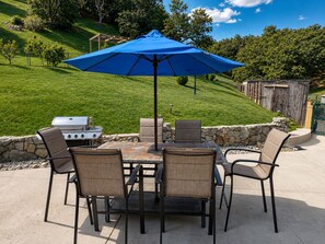 Een terras buiten met een barbecue, tafel, stoelen en een blauwe parasol.