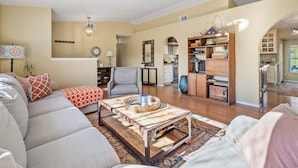 Uma sala de estar ampla com um sofá, poltrona e mesa de centro. Uma cozinha é visível ao fundo, com armários e um espaço para refeições.
