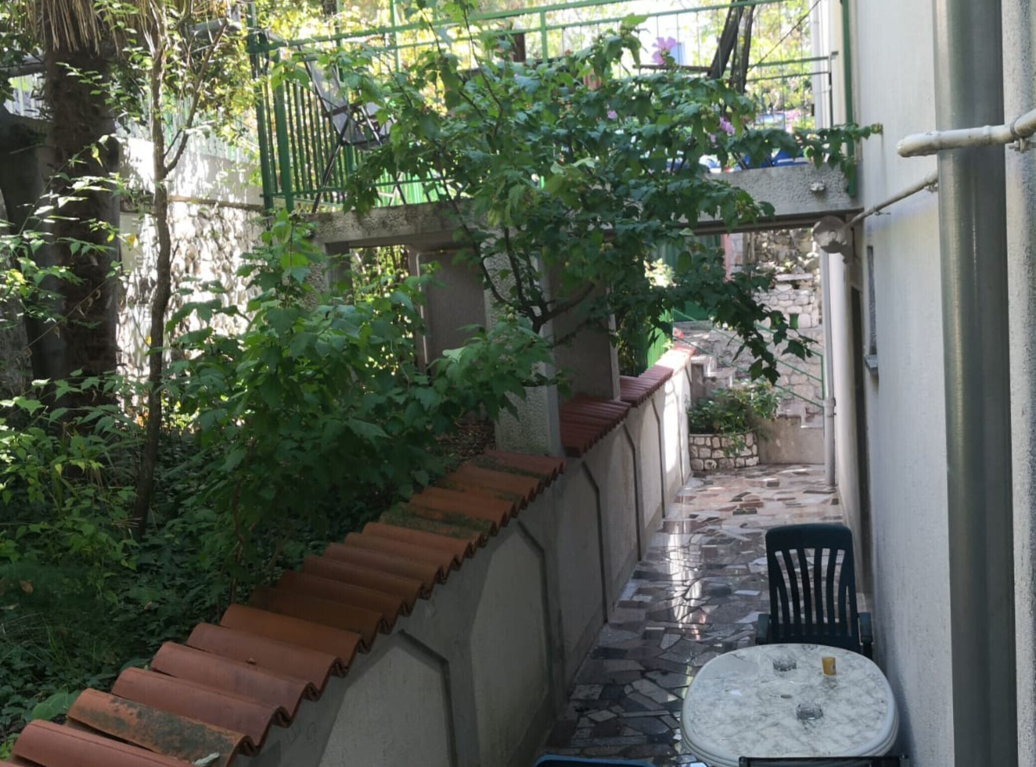 A narrow outdoor corridor with a tiled floor, a metal chair, and a table with a cup.
