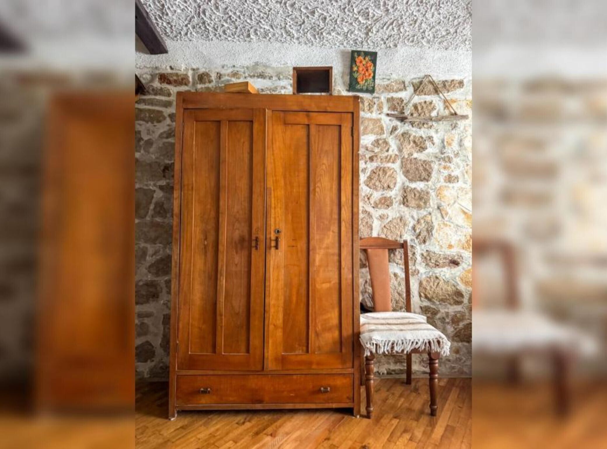 Une armoire en bois, avec un mur en pierre en arrière-plan et une chaise dont l’assise est en osier tressé.