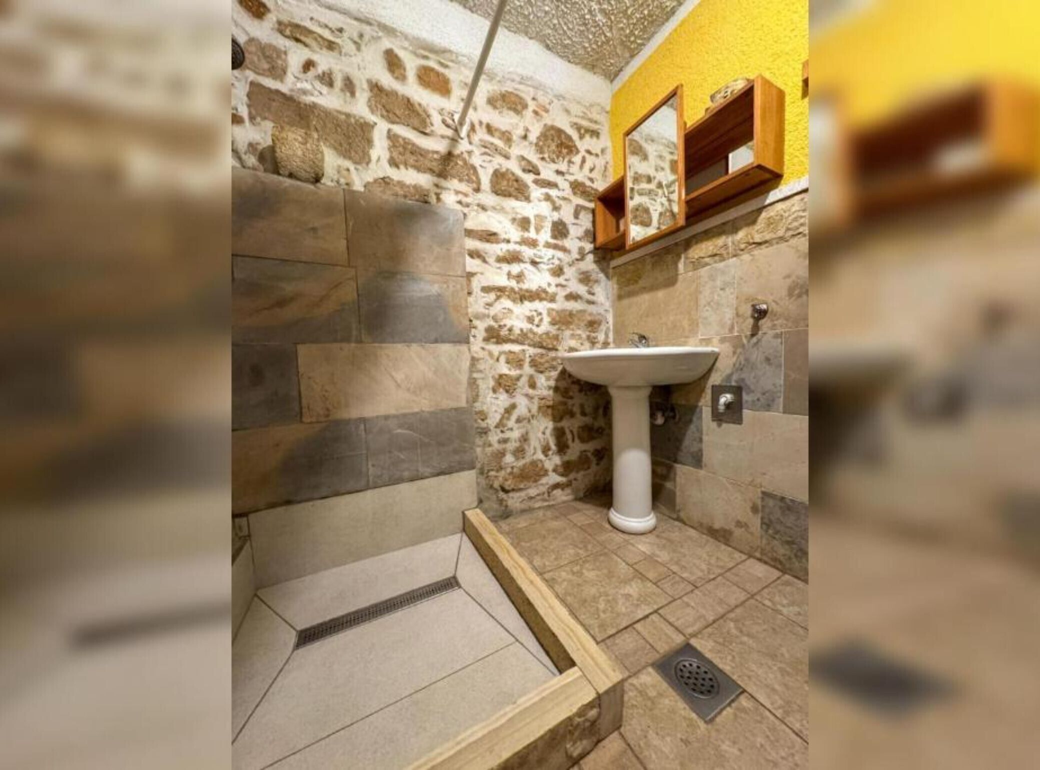 A bathroom with a stone wall, a white pedestal sink, and a wooden shelf above it.