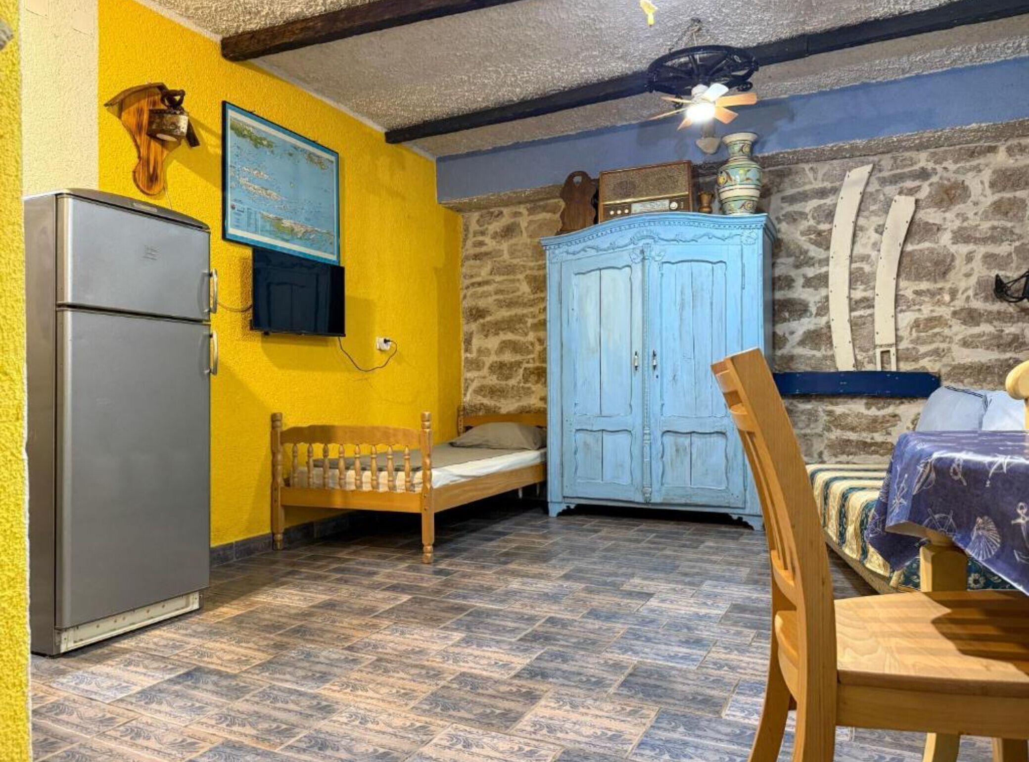 Una habitación con una cama, una silla de madera, una mesa, un armario azul, un mapa en la pared y un refrigerador.