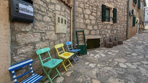 Un bâtiment en pierre à l’aspect rustique, un petit espace extérieur aménagé avec des chaises colorées, et un coffret électrique fixé au mur.