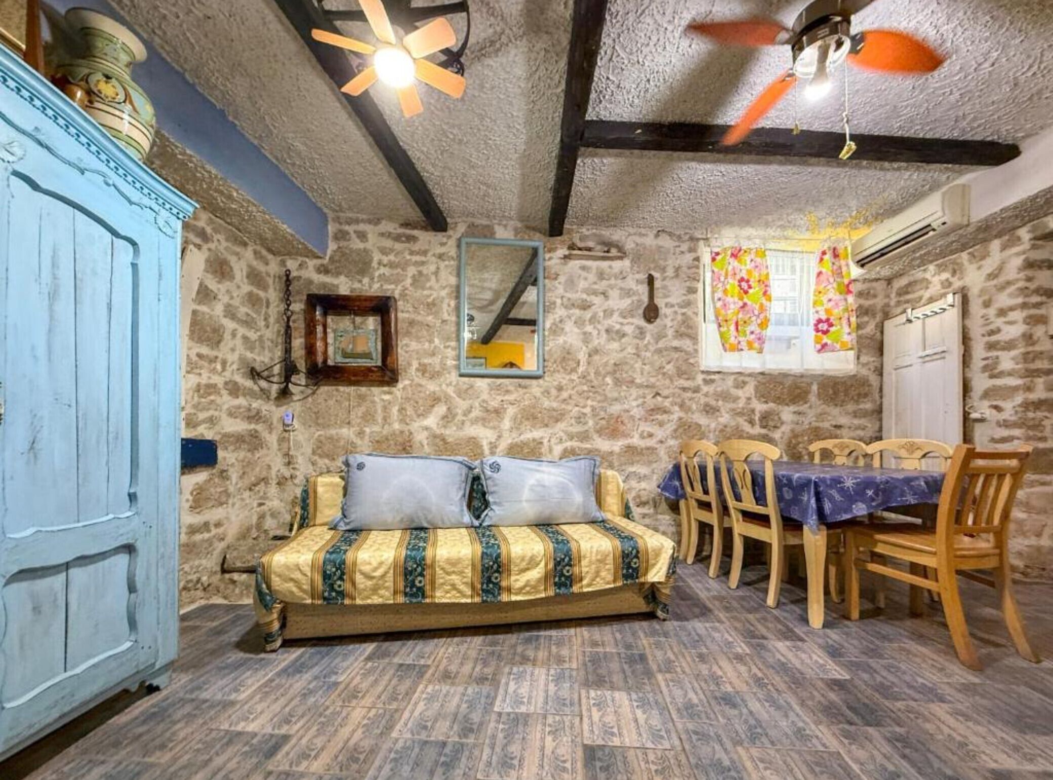 Quarto rústico com parede de pedra, cama de madeira com roupa de cama estampada, mesa de jantar com cadeiras e um ventilador de teto.