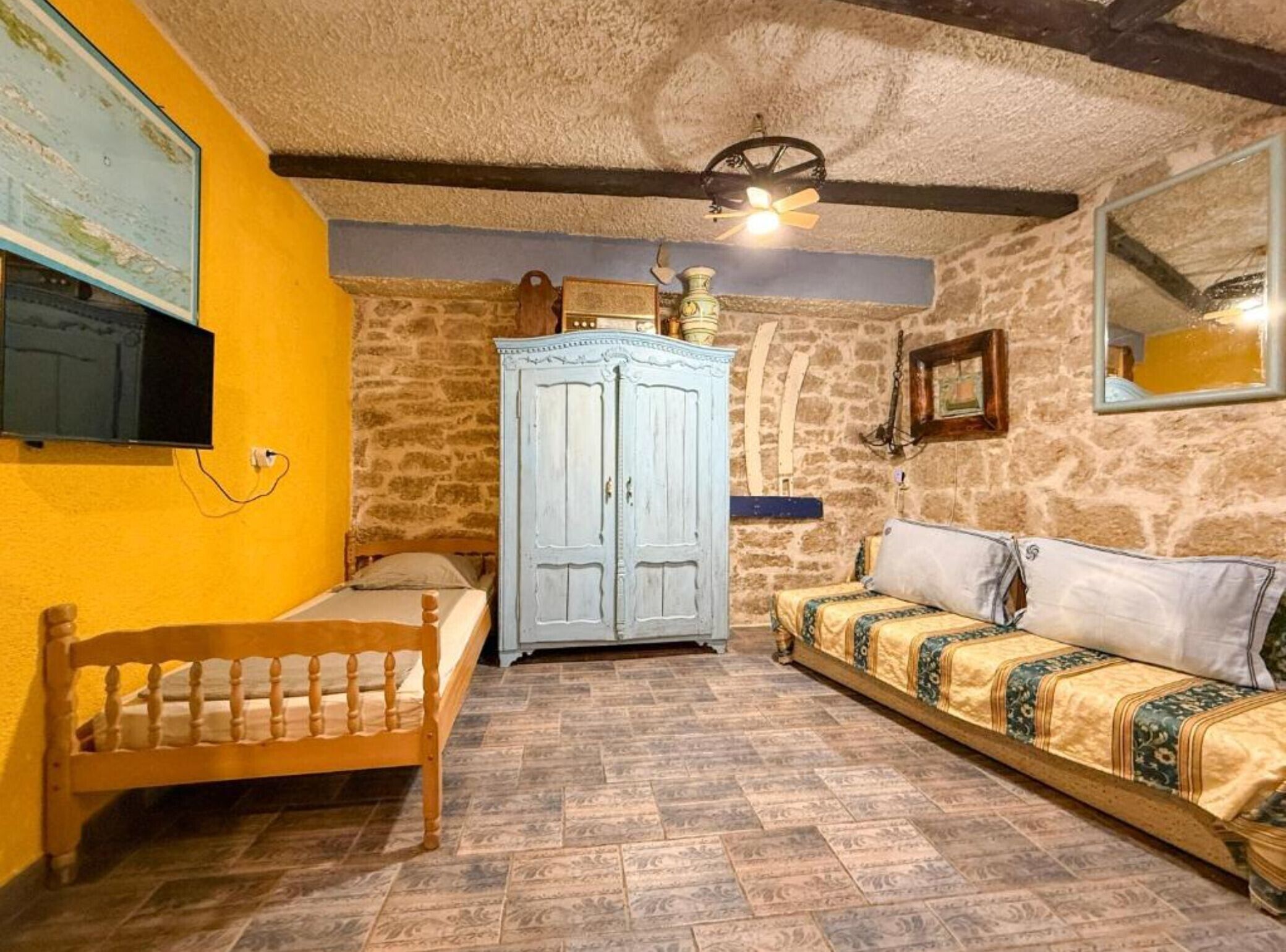 A room with a wooden bed, a blue wardrobe, a stone wall, and a ceiling fan.
