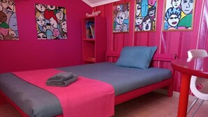 A pink-themed room with a single bed, a small table, and colorful artwork on the walls. - Magic mola (Chalkidiki)