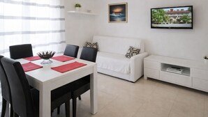 A modern, minimalist living room with a white sofa, a dining table with red placemats, a TV on the wall, and a small shelf with a plant. - Apartments Anita - One bedroom apartment with terrace (Stobrec)