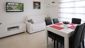 A modern living room with a white sofa, a dining table with chairs, a TV mounted on the wall, and a window with curtains. - Apartments Anita - One bedroom apartment with terrace (Stobrec)