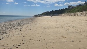 Ein weiter Sandstrand mit vereinzelten Felsen und Fußabdrücken, ein klarer Himmel mit Wolken sowie eine entfernte Küstenlinie mit Gebäuden.