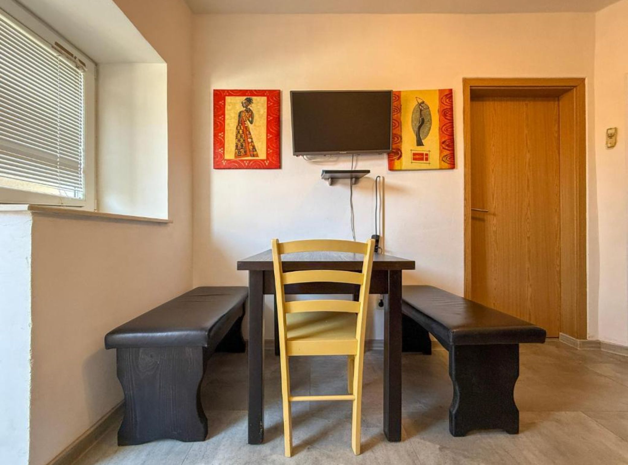 A room with a wooden chair and bench, a flat-screen TV mounted on the wall, and two framed artworks above the TV.