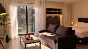 A modern hotel room with a sofa, a bed, a table, and a view of greenery outside. - Alma Rentals Sol Palermo (Buenos Aires)