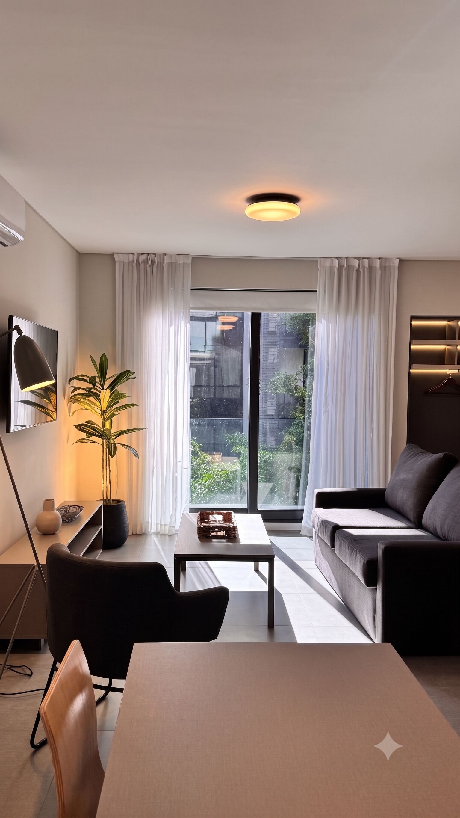 Un salon moderne avec un canapé, une table à manger et un téléviseur. De grandes fenêtres sont ornées de rideaux, et on y trouve une plante en pot ainsi qu’un luminaire suspendu.