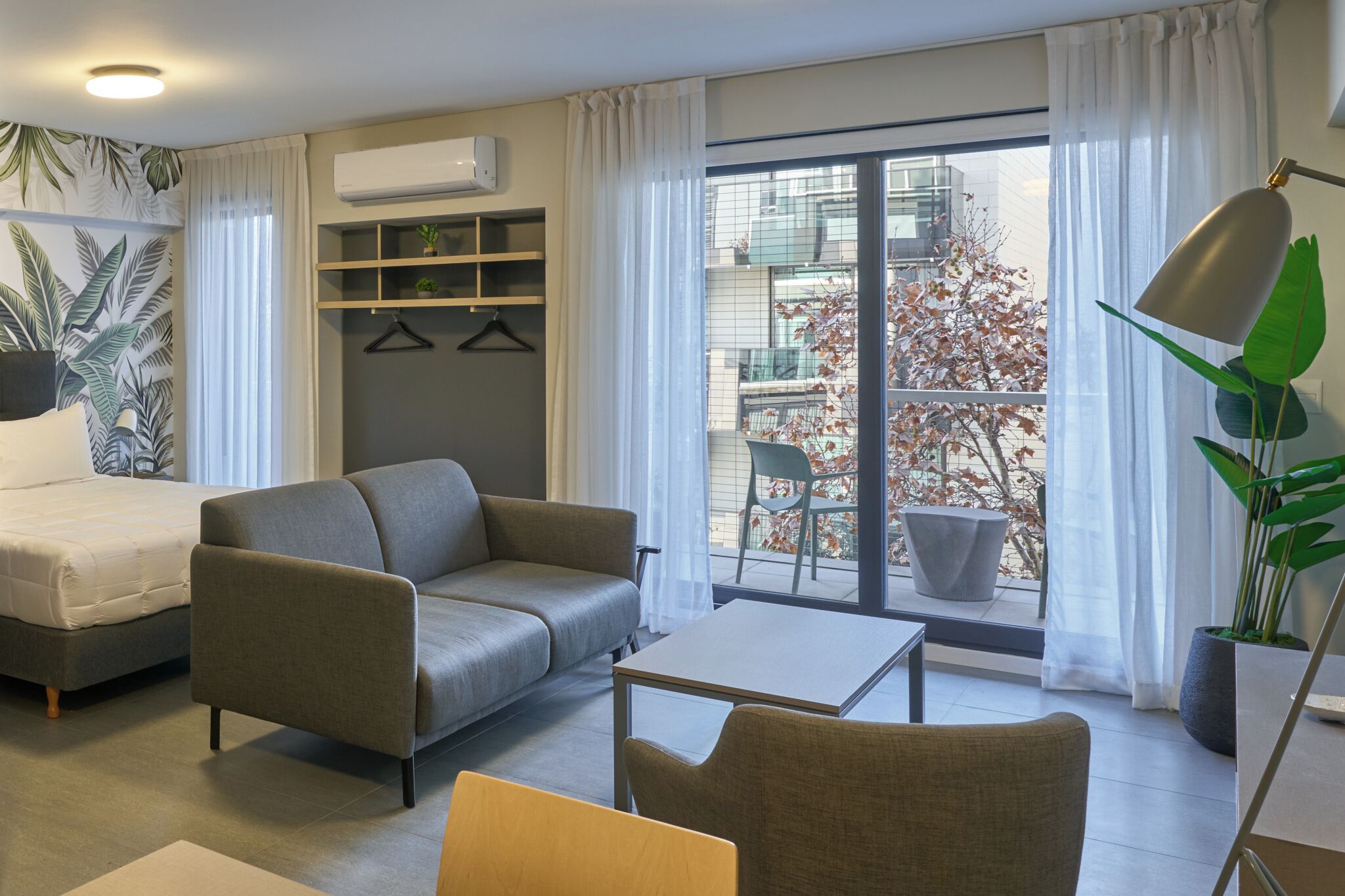 A modern living room with a sofa, a small table, and a chair. There is a balcony with a view of buildings and a tree.