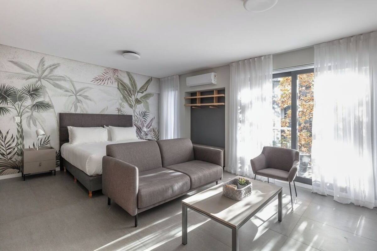 A modern hotel room with a bed, sofa, armchair, and a coffee table.