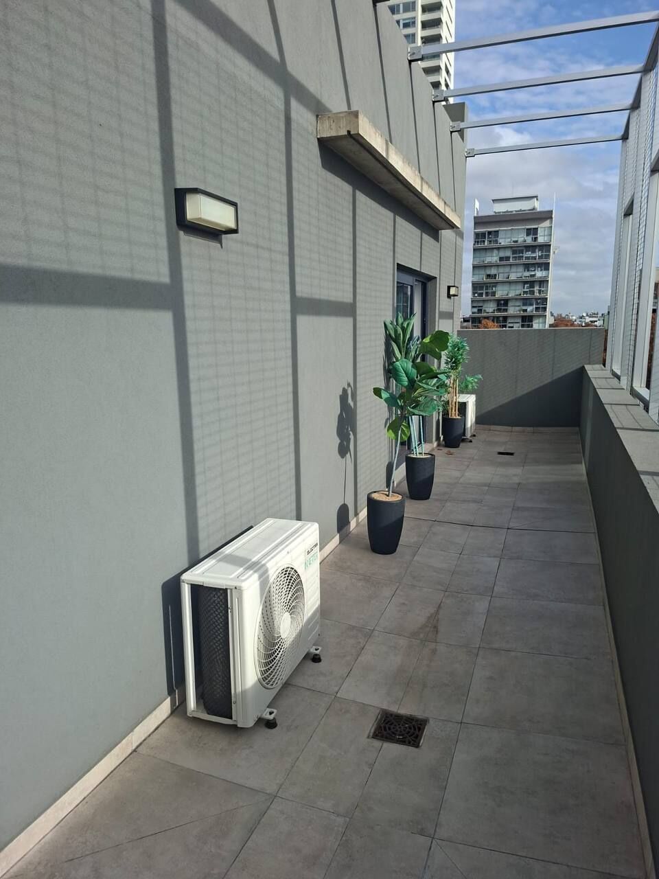 Um corredor no terraço com unidade de ventilação, plantas em vasos e vista para um edifício ao fundo.