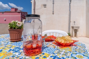 A table with a colorful tablecloth, a glass jar with a black lid, a potted plant, and a bowl of chips. - Apartment 'Suli Mari Amuri' with Private Terrace (Avola)
