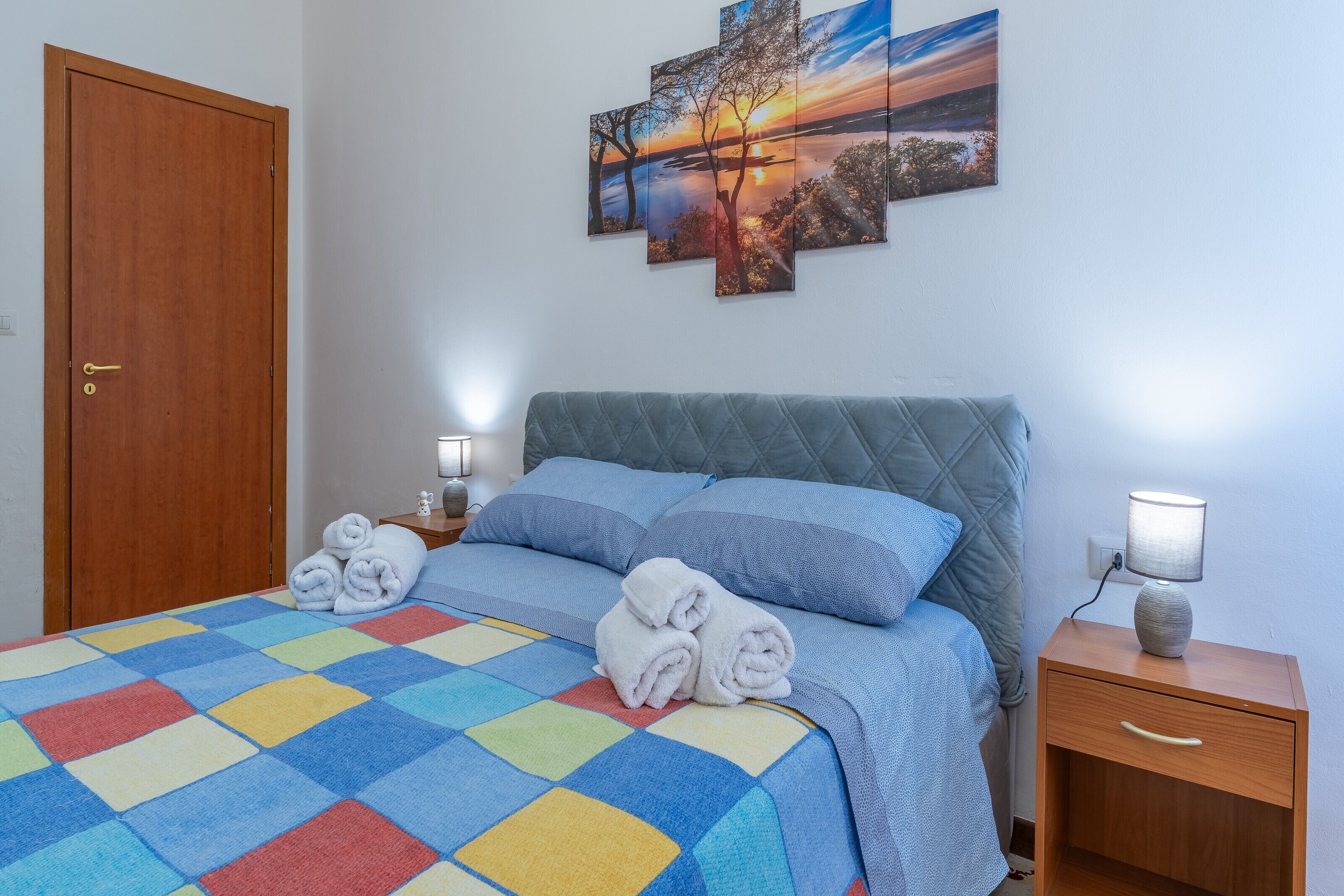 Ein ordentlich bezogenes Bett mit einer farbenfrohen Steudecke, ein Nachttisch aus Holz mit einer Lampe und eine Tür.