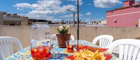 A rooftop terrace with a table set for two, a potted plant, a large jar, and a basket of chips.