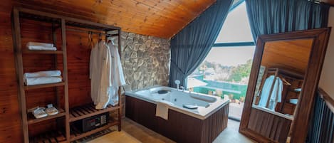 Un baño de cabaña de madera con pared de piedra, estante de madera con toallas, una bañera y vistas por una ventana.