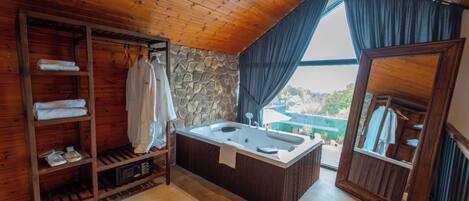 A wooden cabin with a stone wall, a wooden shelf with towels, a bathtub, and a view through a window.