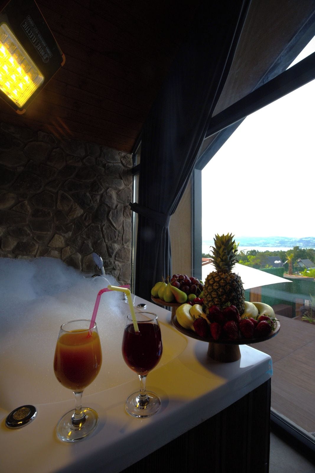 Zona de bar con dos vasos de bebida roja, un frutero y vista al exterior.