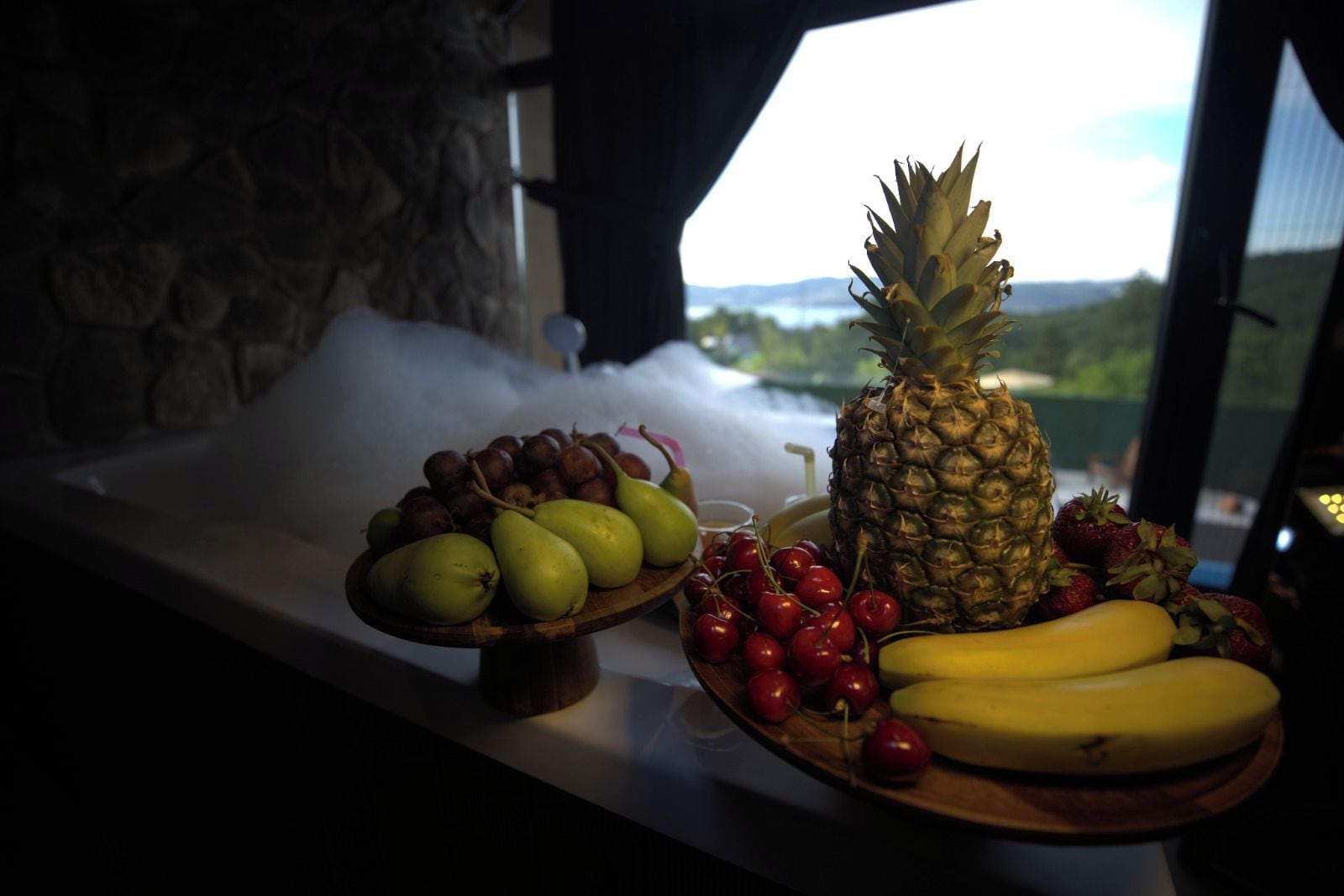 A fruit platter on a table with a view of a landscape through a window.