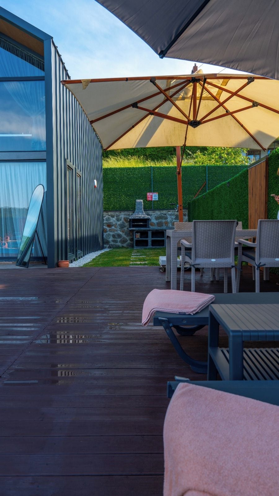 Eine Terrasse im Freien mit Tisch und Stühlen unter einem großen Sonnenschirm, einem Stein-Barbecue und Blick auf einen grünen Heckenstreifen und ein Gebäude.