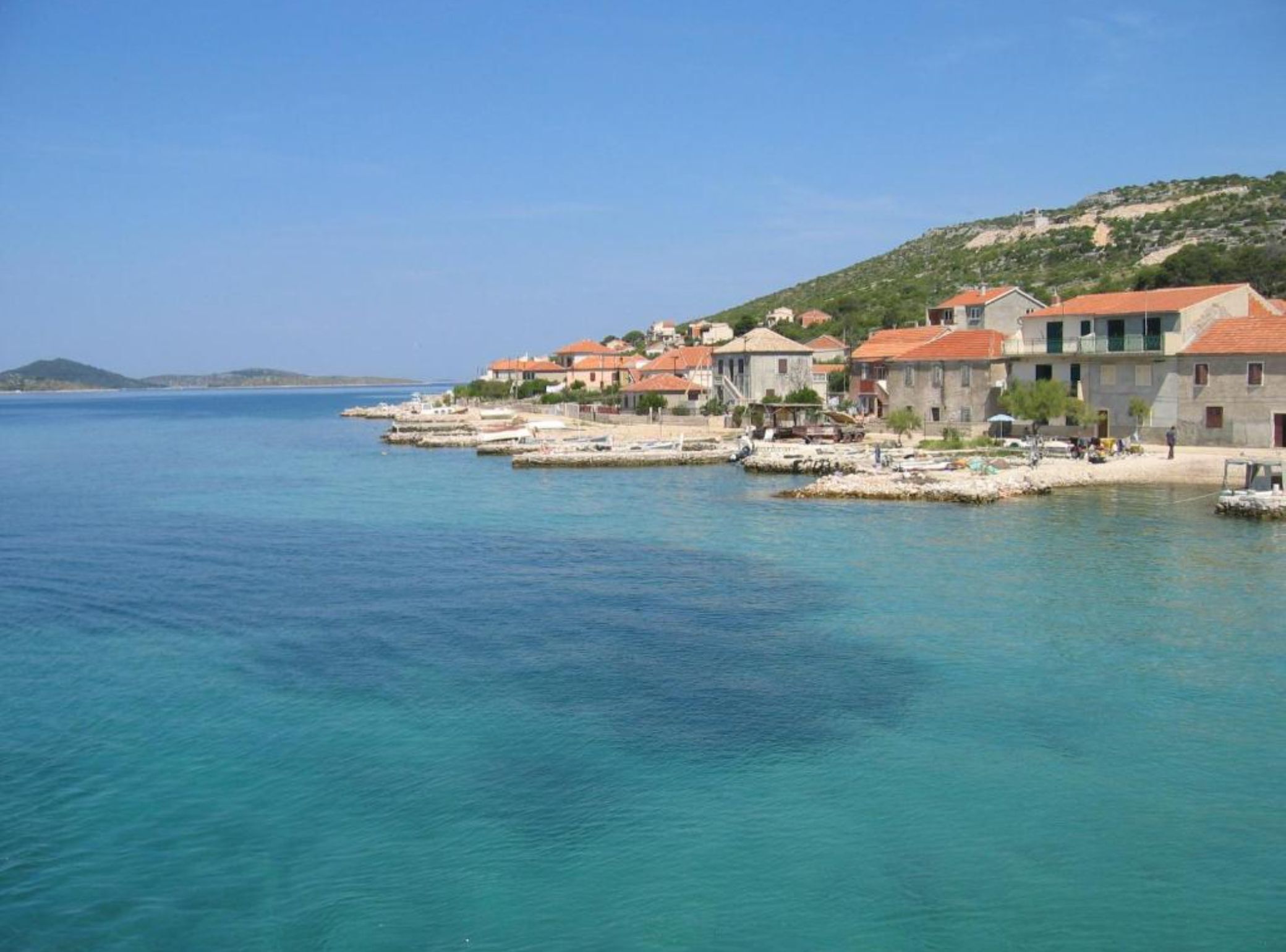 Un village côtier aux bâtiments en pierre, bordé par une mer d’un bleu limpide, et dominé par des collines.