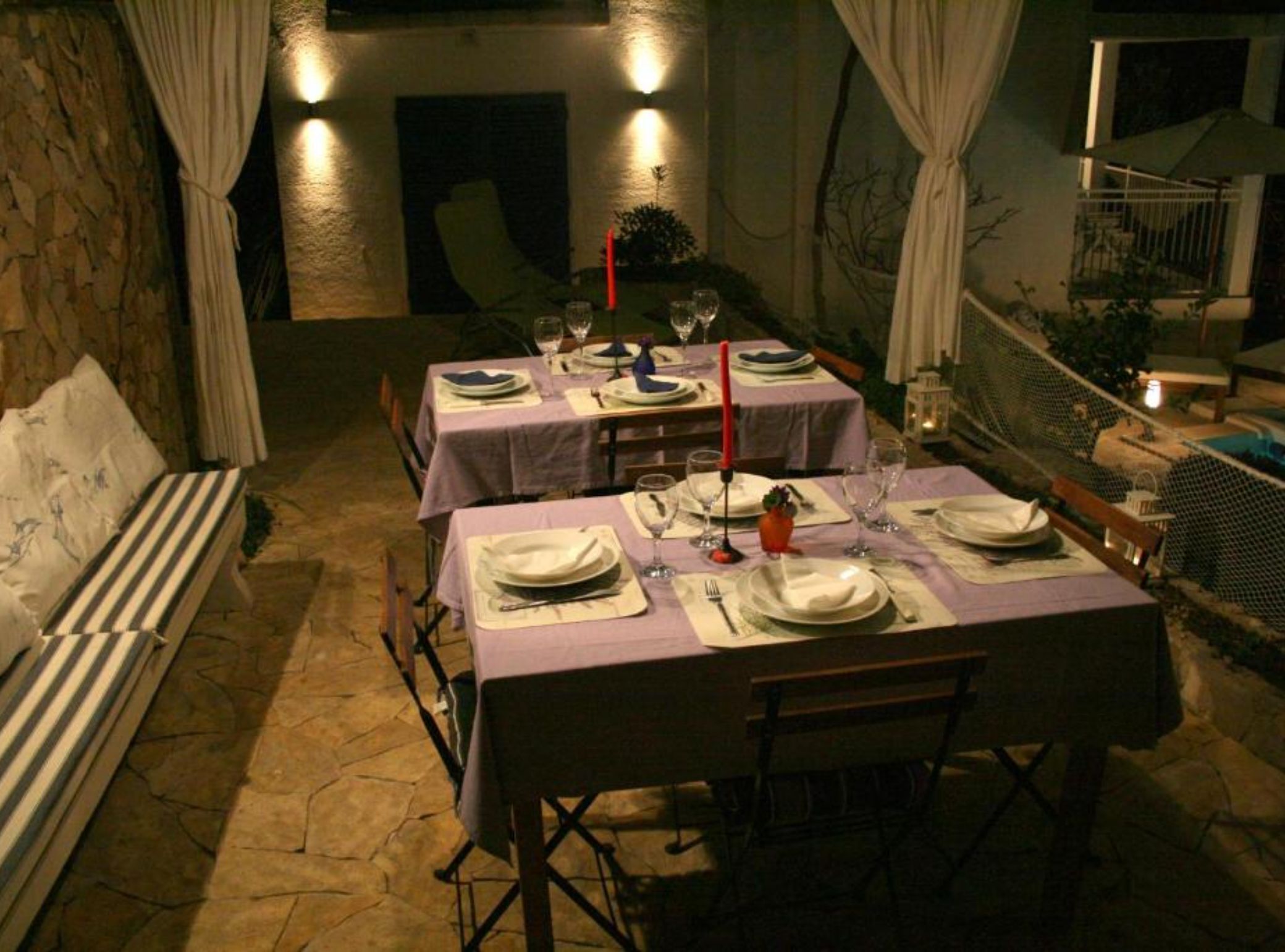 Un espacio de comedor al aire libre con suelo de piedra, mesas dispuestas con manteles blancos y velas.