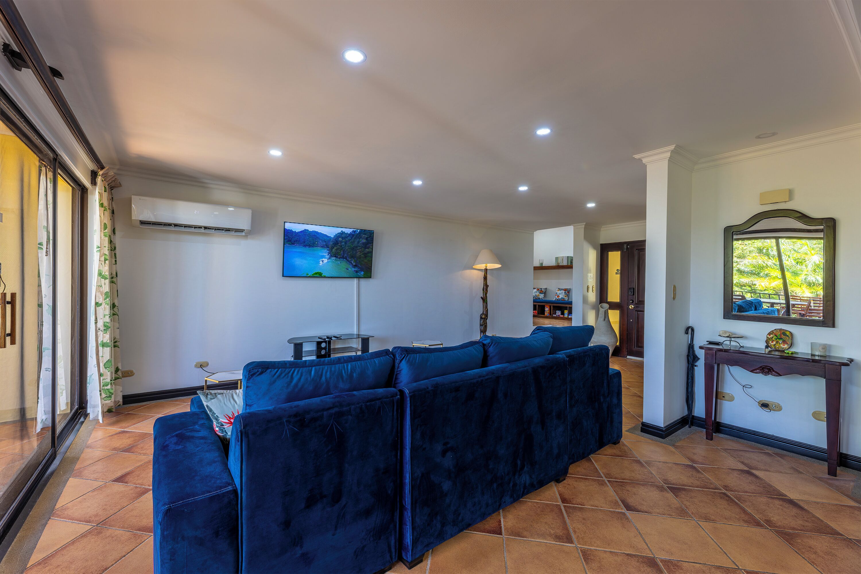 A living room with a sectional sofa, an air conditioning unit, a television, and a dining area with a table and chairs.