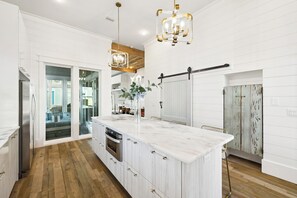 A modern kitchen with a large island, wooden flooring, white cabinetry, and pendant lighting. - Home w/ Carriage, Private Pool and Peloton (Santa Rosa Beach)
