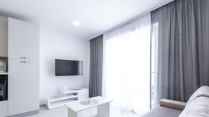 Een moderne woonkamer met een witte bank, een witte salontafel, een flatscreen-tv aan de muur en een keukenhoek met kastjes en een magnetron.