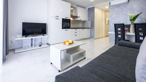 Een moderne woonkamer met een flatscreen televisie, een bijzettafeltje met fruit, een bank en een keukenhoek met kasten en apparatuur.
