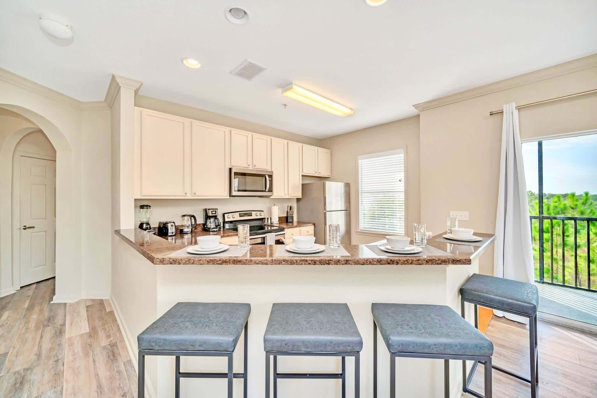 A modern kitchen with a central island, bar stools, and a view of greenery through the window.