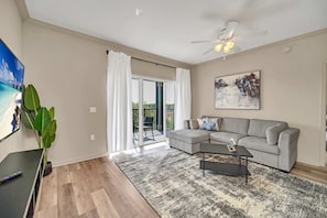A living room with a grey sofa, a black coffee table, a ceiling fan, and a large window with white curtains. - Upstay Modern 2BR Condo Near Disney (Winter Garden)