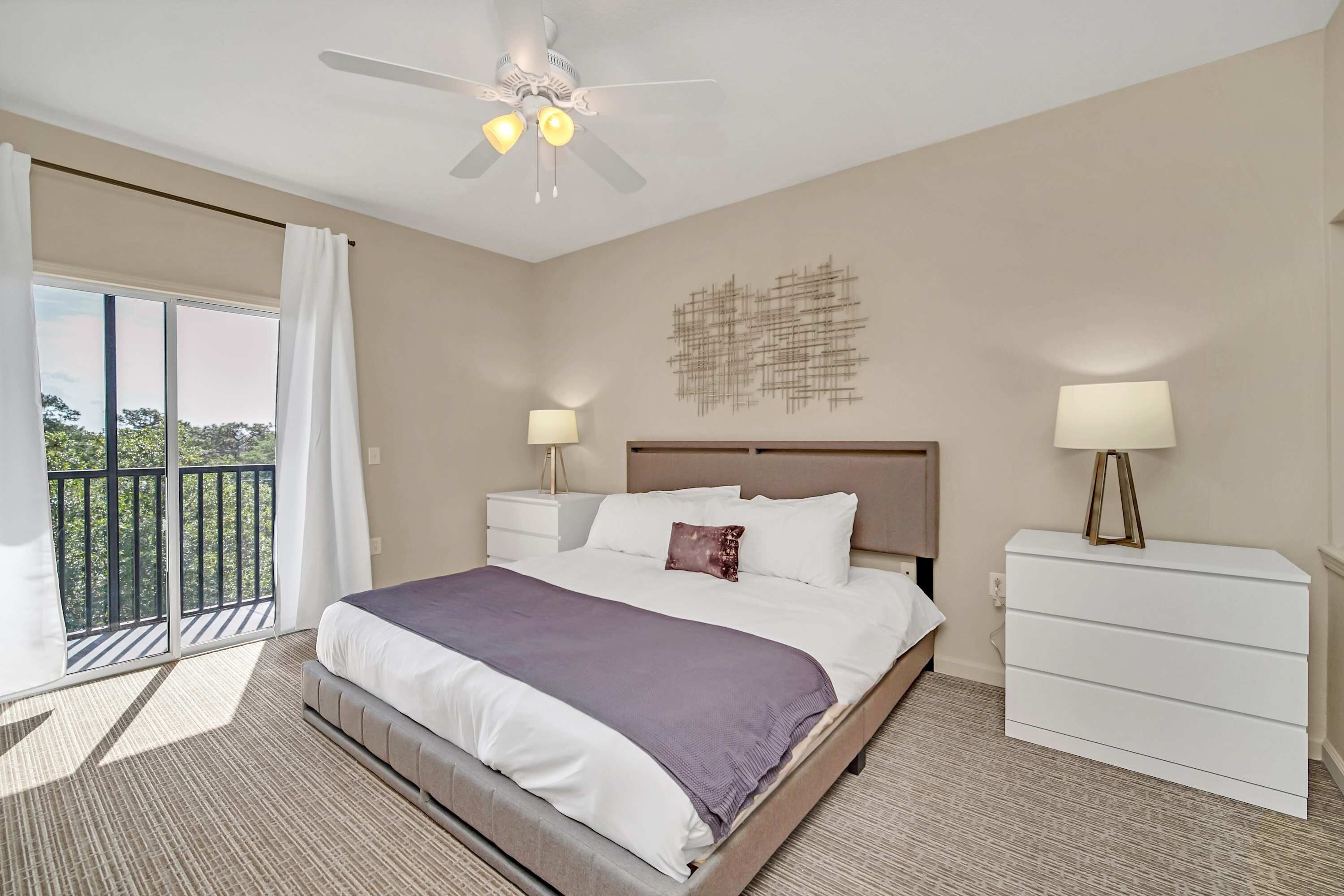 A hotel room with a bed, two bedside tables, a ceiling fan, a balcony with curtains, and a wall-mounted abstract art piece.