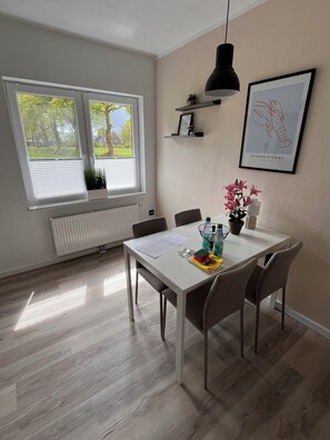 Un comedor con una mesa blanca, cuatro sillas, una lámpara colgante negra, un radiador, una ventana con persianas y un cuadro en la pared.