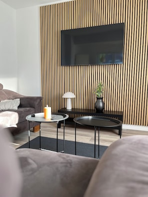 A modern living room with a sofa, two round tables, a lamp, a TV, and a wooden wall panel. - Since April 2026: Modern, charming vacation home, large Terrace with a great view! (Aurich)