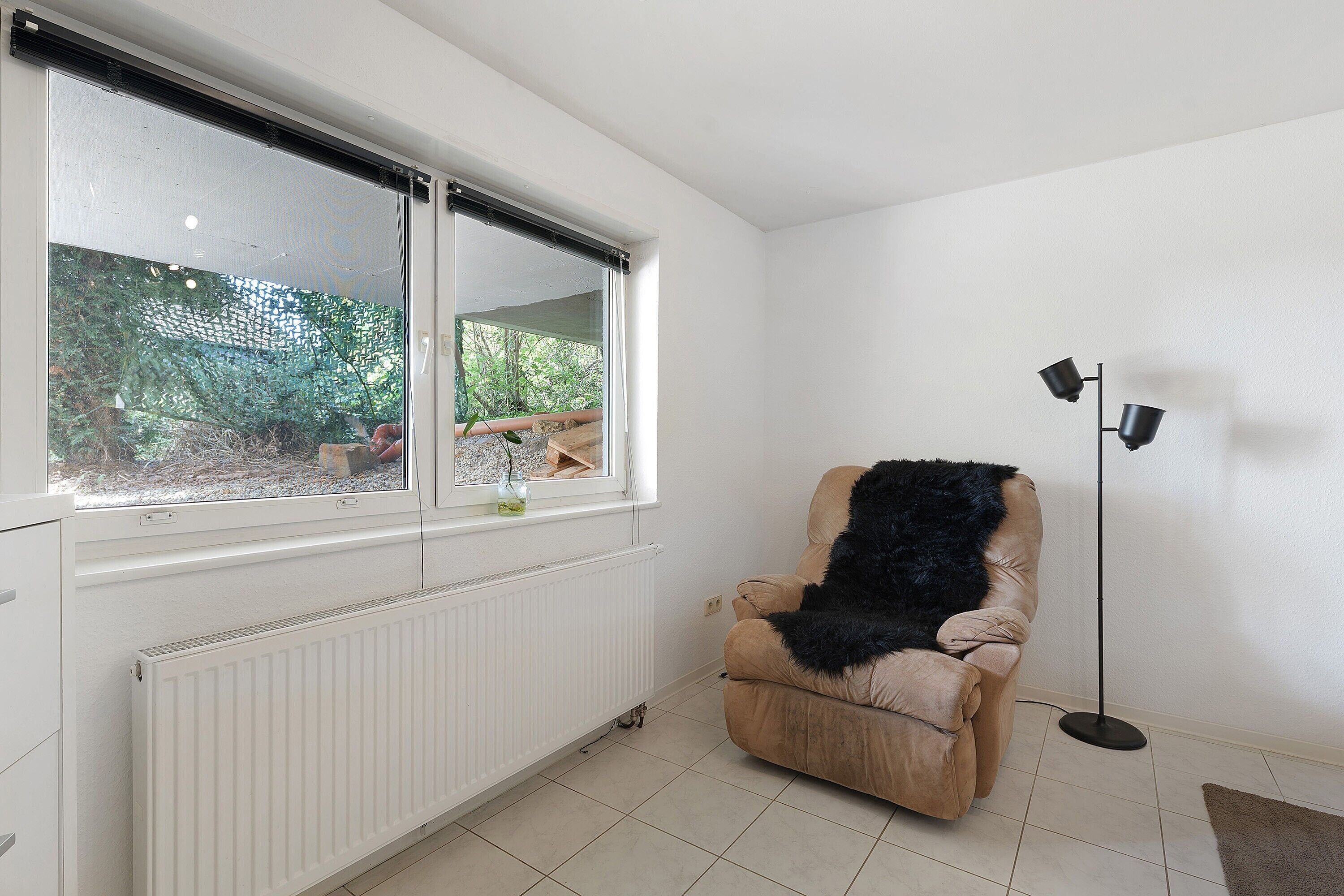Una habitación con un radiador, un ventanal con vistas a follaje, una silla con un plaid negro y una lámpara de pie.